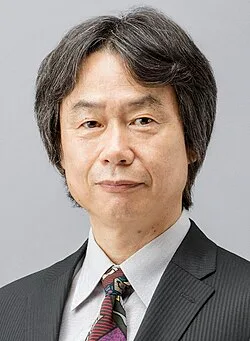 Shigeru Miyamoto, a Japanese man wearing a black coat and white shirt with red, yellow, and blue Pikmin characters